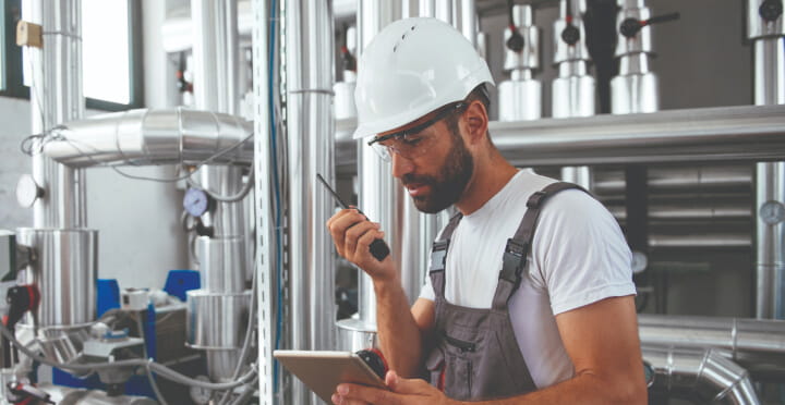 An engineer holds a tablet and walkie-talkie in boiler room.