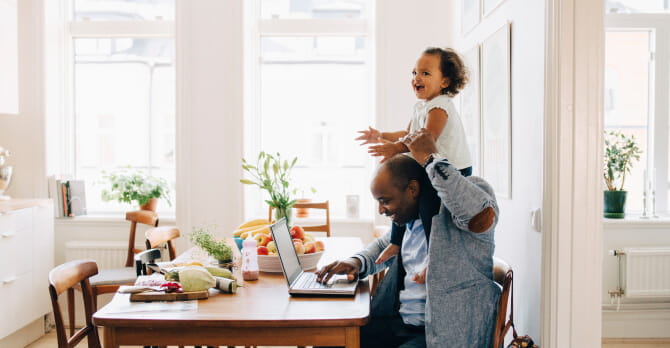 Father holding young daughter on his shoulders while working on laptop at kitchen table
