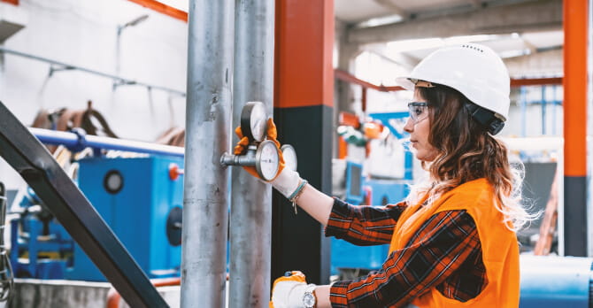 Woman in hard hat and protective googles inspecting valves in factory
