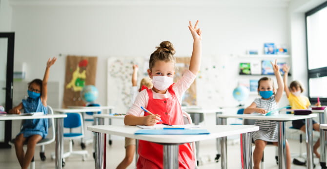 Young children with protective face masks on sitting at desks in a classroom, raising hands
