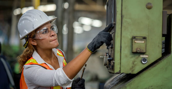 Woman in hard hat and safety glasses leans in to inspect heavy equipment.