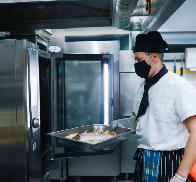 A chef with a protective face mask putting food in an industrial oven