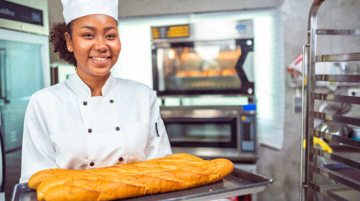 A young chef holds a tray with bread while standing in a modern commercial kitchen.