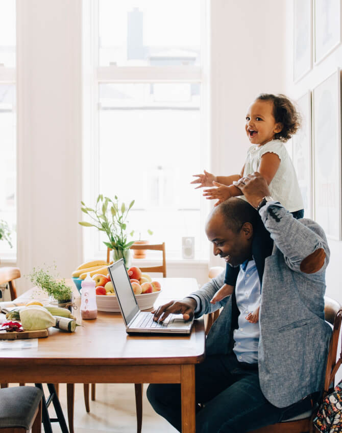 Father holding young daughter on his shoulders while working on laptop at kitchen table