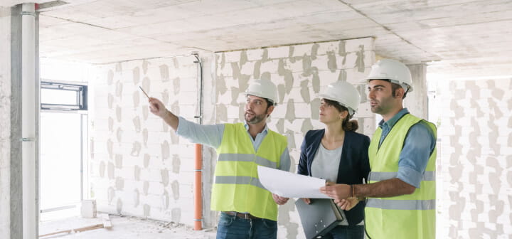 Three developers examine the layout of a building under construction, in relation to a blueprint.