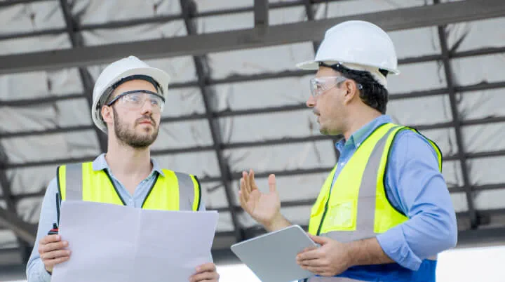 Two workers wearing safety equipment while having a discussion at a construction site. One worker holds a blueprint, the other holds an Ipad. 
