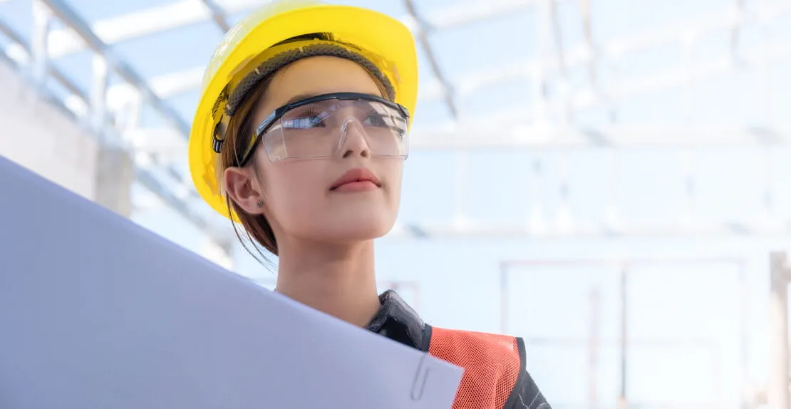 Engineer on a construction site holding a blueprint  while wearing a safety helmet and goggles.