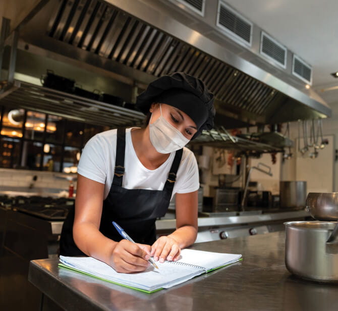 Smiling woman in restaurant kitchen wearing a protective face mask and writing in a spiral notebook