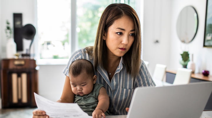 A parent sits in front of a laptop while a baby sits on their lap. Paperwork is laid across the desk. 