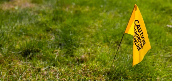 A bright-yellow safety flag stuck into a green lawn. Partially hidden text on the flag reads “Caution: Underground pipeline.”