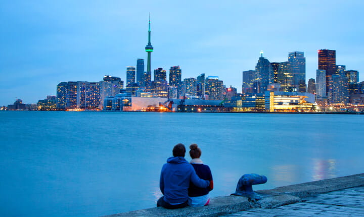 Photo of a couple sitting close together on a pier, admiring the downtown Toronto skyline illuminated at night.