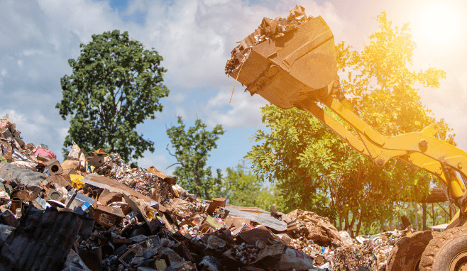 A tractor scoops landfill waste from a large pile of refuse.