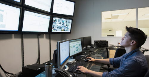 Employee in a control room working on a computer and looking at multiple screens on a wall.
