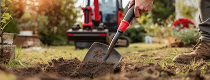 A person digging with shovel in garden soil beside a small excavator at a residential backyard 