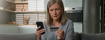 A woman sitting on a couch with phone in one hand and looking at a credit card in the other hand with worried face expressions