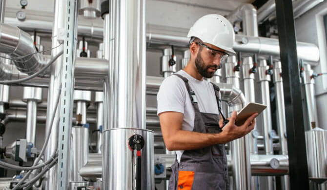 Man wearing hard hat and protective goggles looking at tablet in boiler room