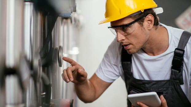 Man in hard hat and safety goggles inspecting natural gas processing machine and holding tablet