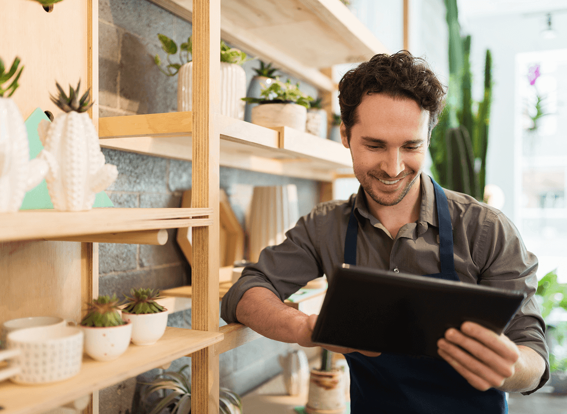 A shop owner in an apron smiles while using a digital tablet in a store filled with potted plants and decorative items on wooden shelves.
