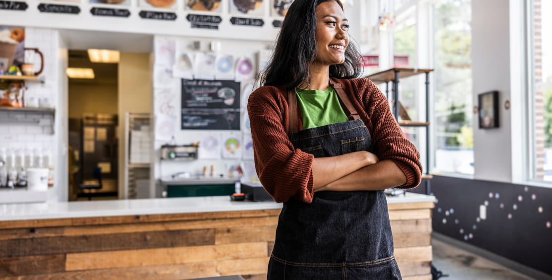 A smiling cafe owner in an apron stands in front of the counter in their shop.
