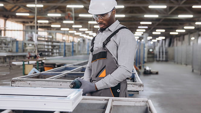 A worker in a hard hat and protective overalls works with metal frames in a large industrial warehouse. The setting is spacious, with overhead lights and rows of machinery and materials in the background.