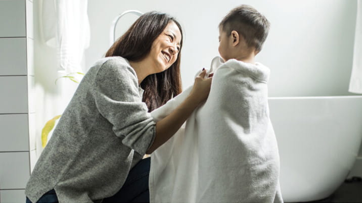 A parent kneels in a modern bathroom and smiles as she wraps her young child in a white towel. The child stands near a freestanding bathtub, and the setting is clean, cozy and softly lit.