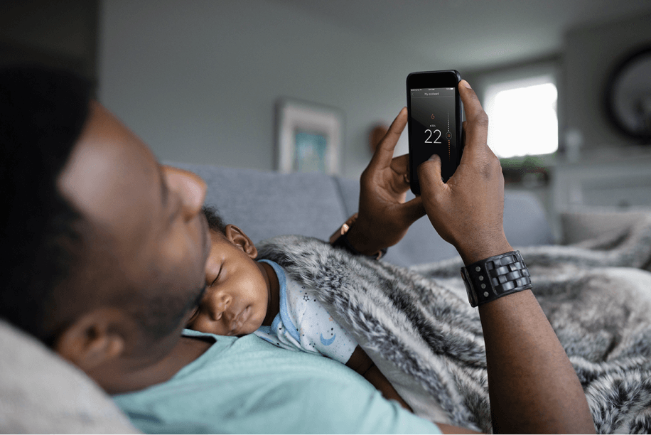 Father lying in bed, adjusting thermostat temperature on phone, while child is sleeping on his chest